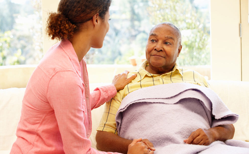 A younger woman sits beside an older man who is covered with a purple blanket, gently placing a hand on his shoulder and holding his hand, offering comfort and support in a bright, sunlit room.