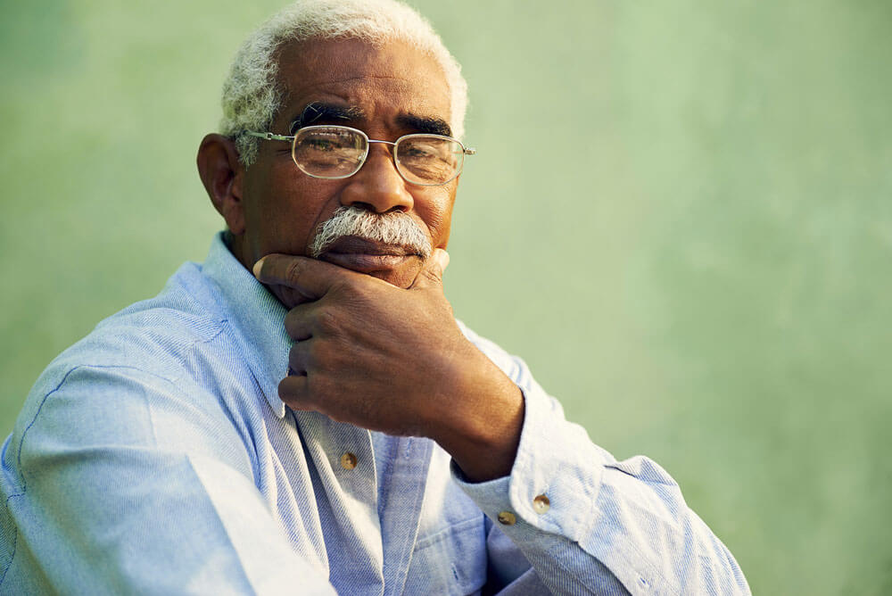 Older man with gray hair and mustache wearing glasses and a light blue shirt, resting his chin on his hand and looking thoughtfully at the camera, with a green background.