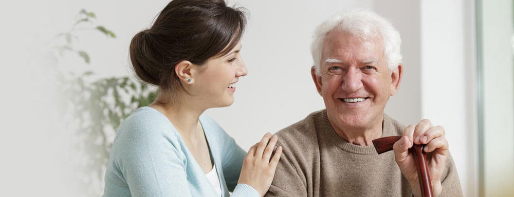 A young woman smiles and talks with an elderly man who is sitting and holding a walking cane, both appearing happy and relaxed in a bright indoor setting.