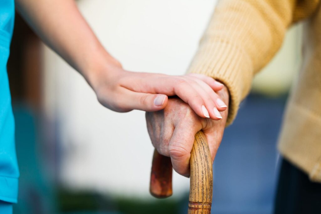 A younger person’s hand rests gently on top of an older persons hand, which is holding a wooden cane. The image conveys support and care between generations.