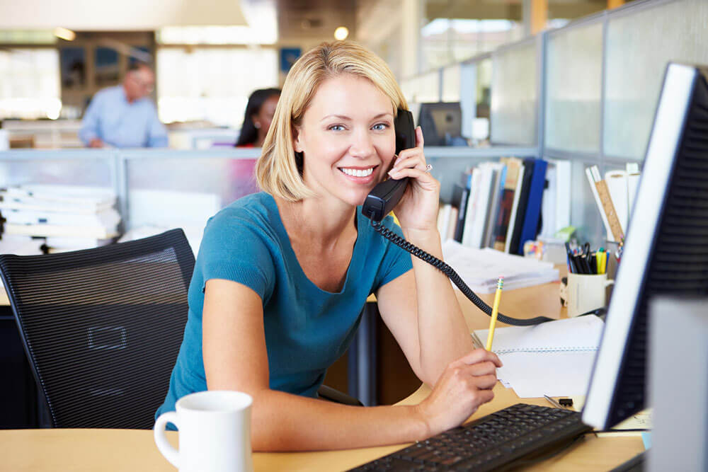 A smiling woman with short blonde hair sits at a desk in an office, holding a phone to her ear and a pencil in her other hand. She faces a computer monitor with a white mug in front of her.