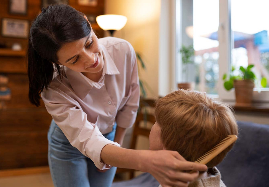 A woman smiles while brushing a young boys hair in a cozy living room with warm lighting and potted plants near the window.