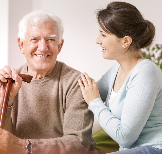 Smiling elderly man with white hair holds a cane while sitting next to a young woman who is gently touching his shoulder and smiling at him. They are indoors, and both look happy and relaxed.