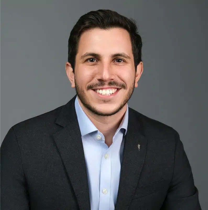 A man with short dark hair and a trimmed beard smiles at the camera. He is wearing a dark blazer over a light blue button-up shirt, and is posed against a plain gray background.