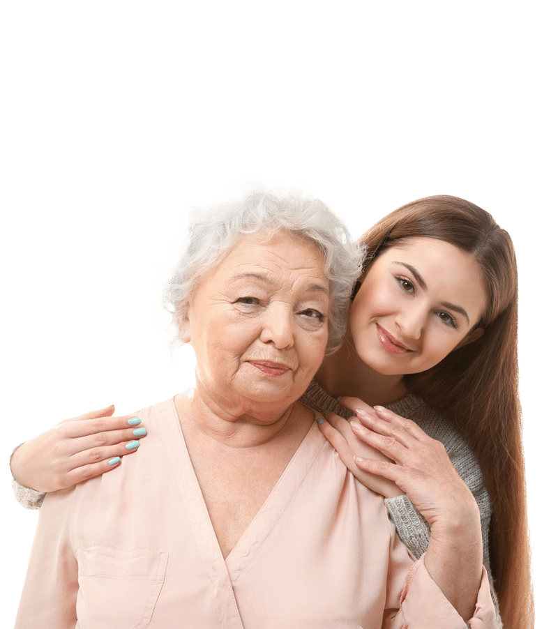 A young woman with long brown hair stands behind an elderly woman with short gray hair, gently hugging her shoulders. Both are smiling softly against a plain white background.