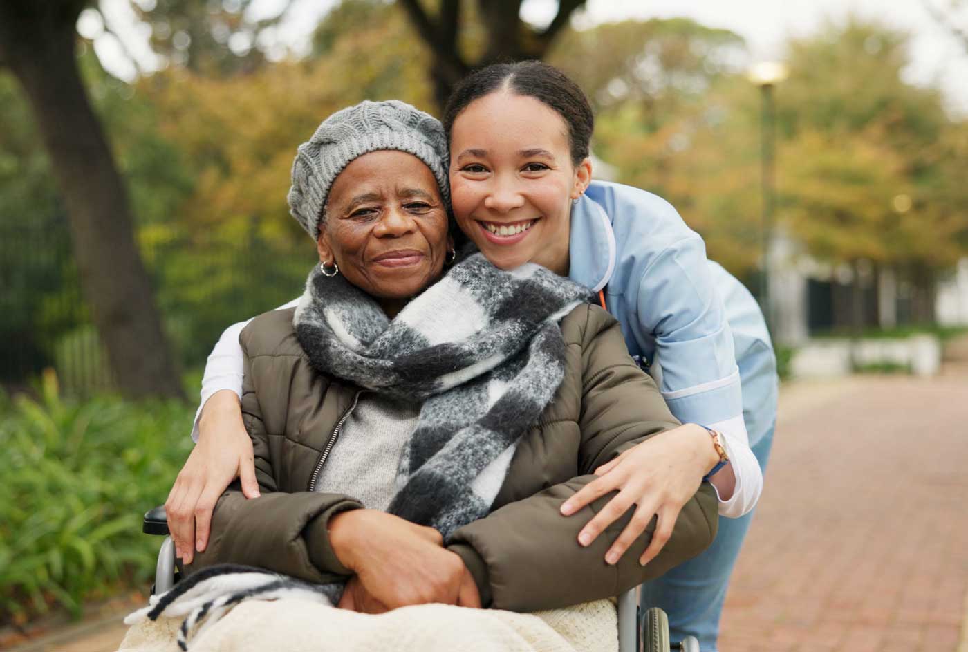 A certified young caregiver in a blue uniform hugs and smiles with an elderly woman in a wheelchair, who is bundled in a hat, scarf, and jacket. They are outside on a path with greenery and trees in the background.