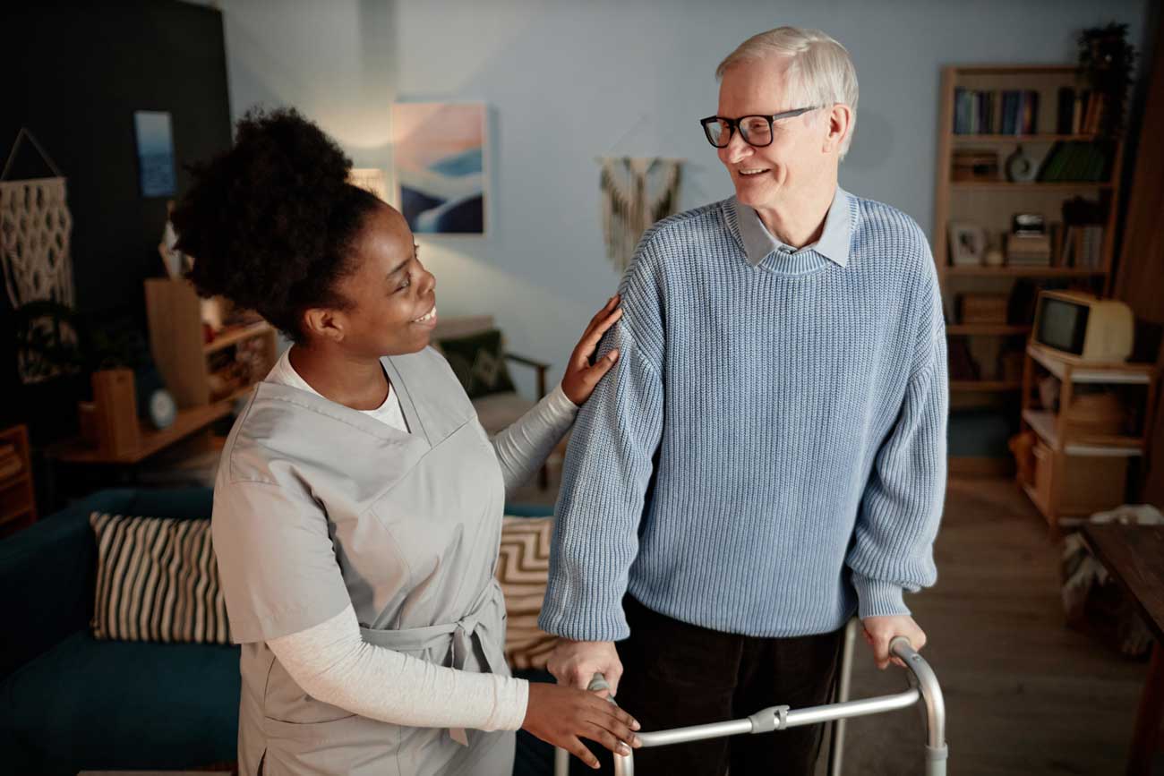 female-caregiver-talking-elderly-man-walking-with-frame-while-helping-patient-around-house