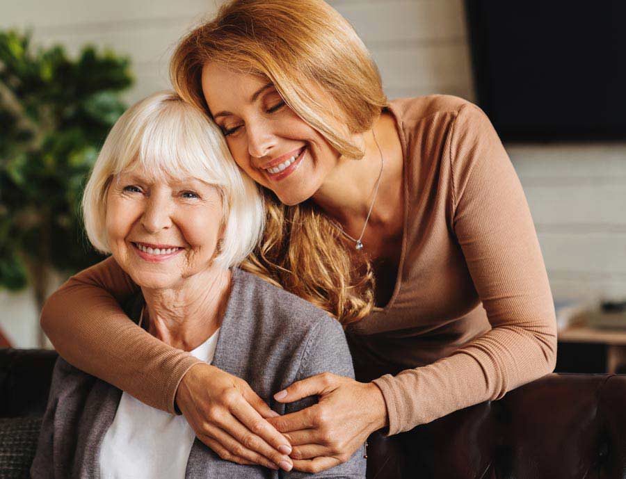 A smiling older woman with short white hair sits while a younger woman with long blonde hair hugs her from behind, both looking happy and relaxed in a cozy indoor setting.