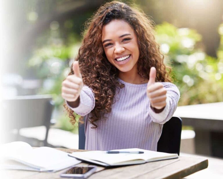 A young woman with curly hair sits at an outdoor table, smiling and giving two thumbs up. Open notebooks and a smartphone are on the table in front of her, with greenery blurred in the background, celebrating her certified achievement.