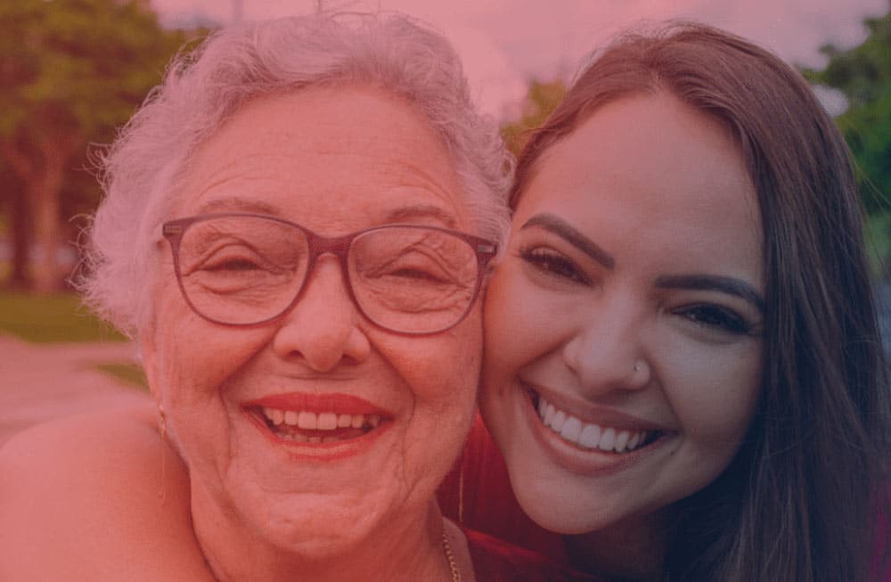 An older woman with glasses and gray hair smiles joyfully next to a younger woman with long dark hair; both are close together, outdoors, and looking at the camera.