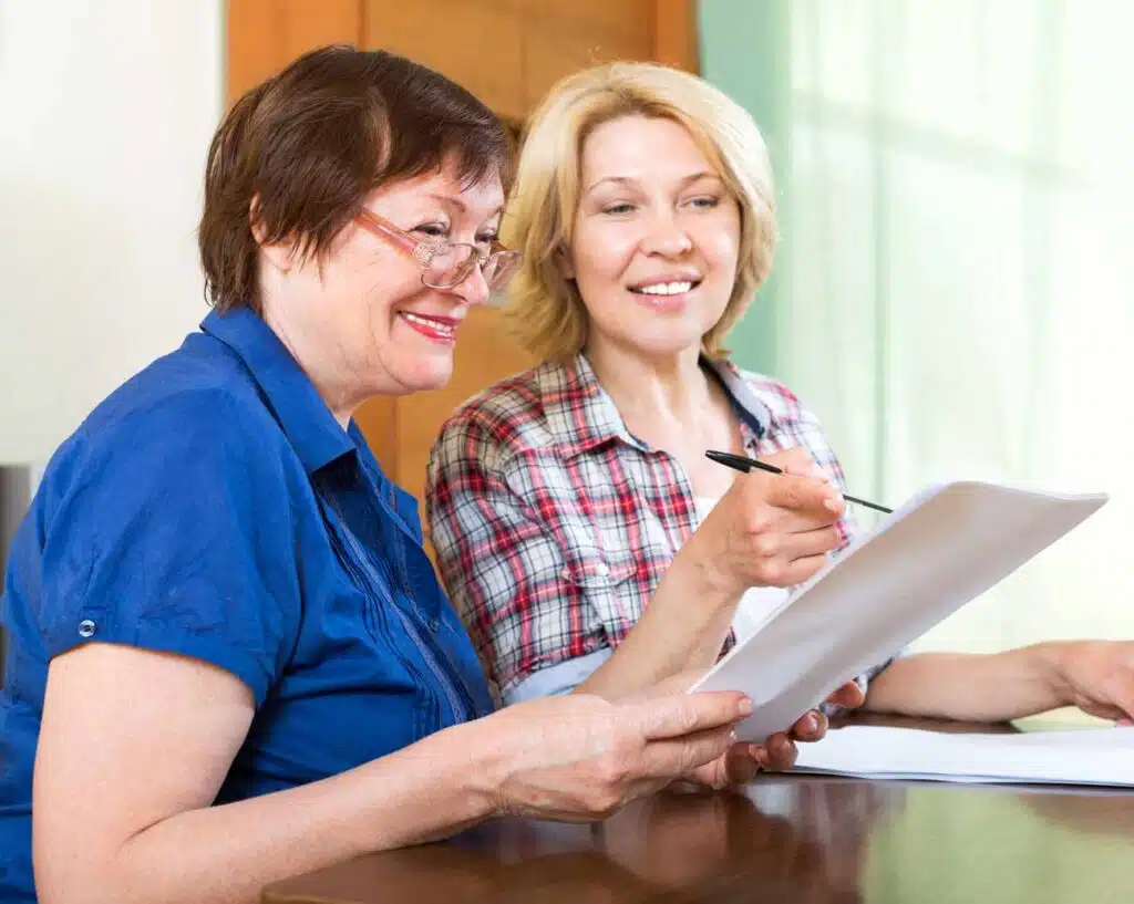 Two middle-aged women sit at a table, smiling and looking at certified documents together. One woman points at the papers with a pen while the other holds them, both appearing engaged and happy.