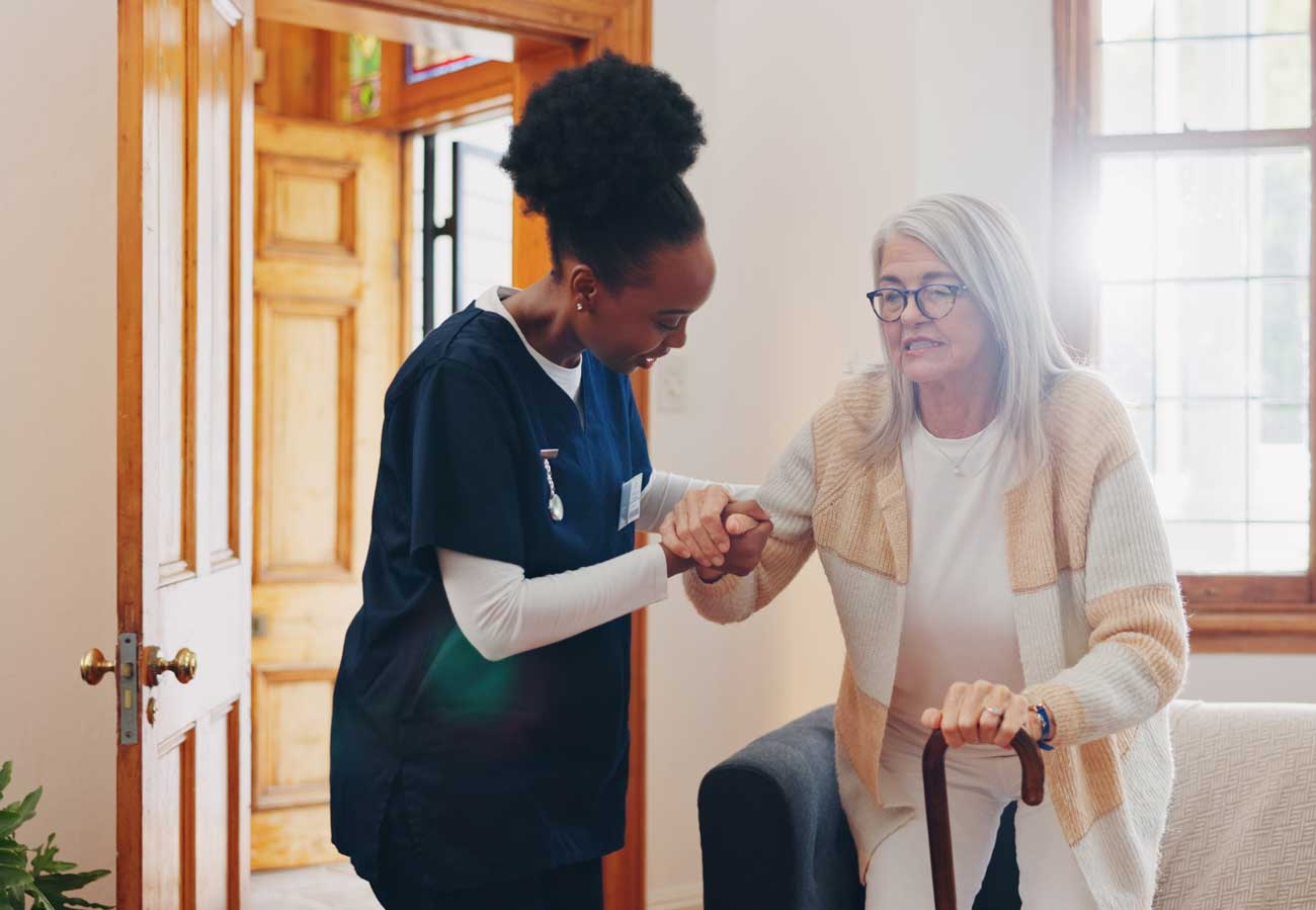A nurse helps an older woman with gray hair and glasses stand up from a sofa. The woman holds a cane, and sunlight streams through the windows in the cozy room.