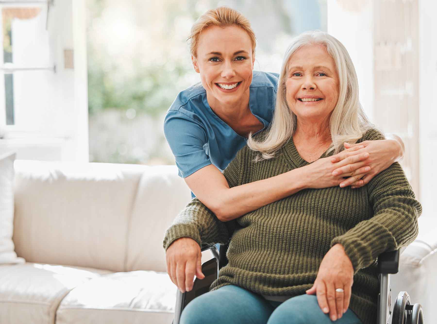 A caring caregiver in blue scrubs hugs an older woman with gray hair, who sits in a wheelchair and wears a green sweater. They are indoors in a bright, comfortable living room.