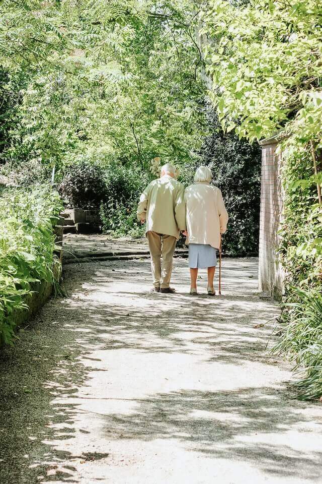 An elderly couple walks arm in arm down a sunlit, tree-lined path. The woman uses a cane, and both are dressed in light clothing. Lush green foliage surrounds them, creating a peaceful, serene atmosphere.