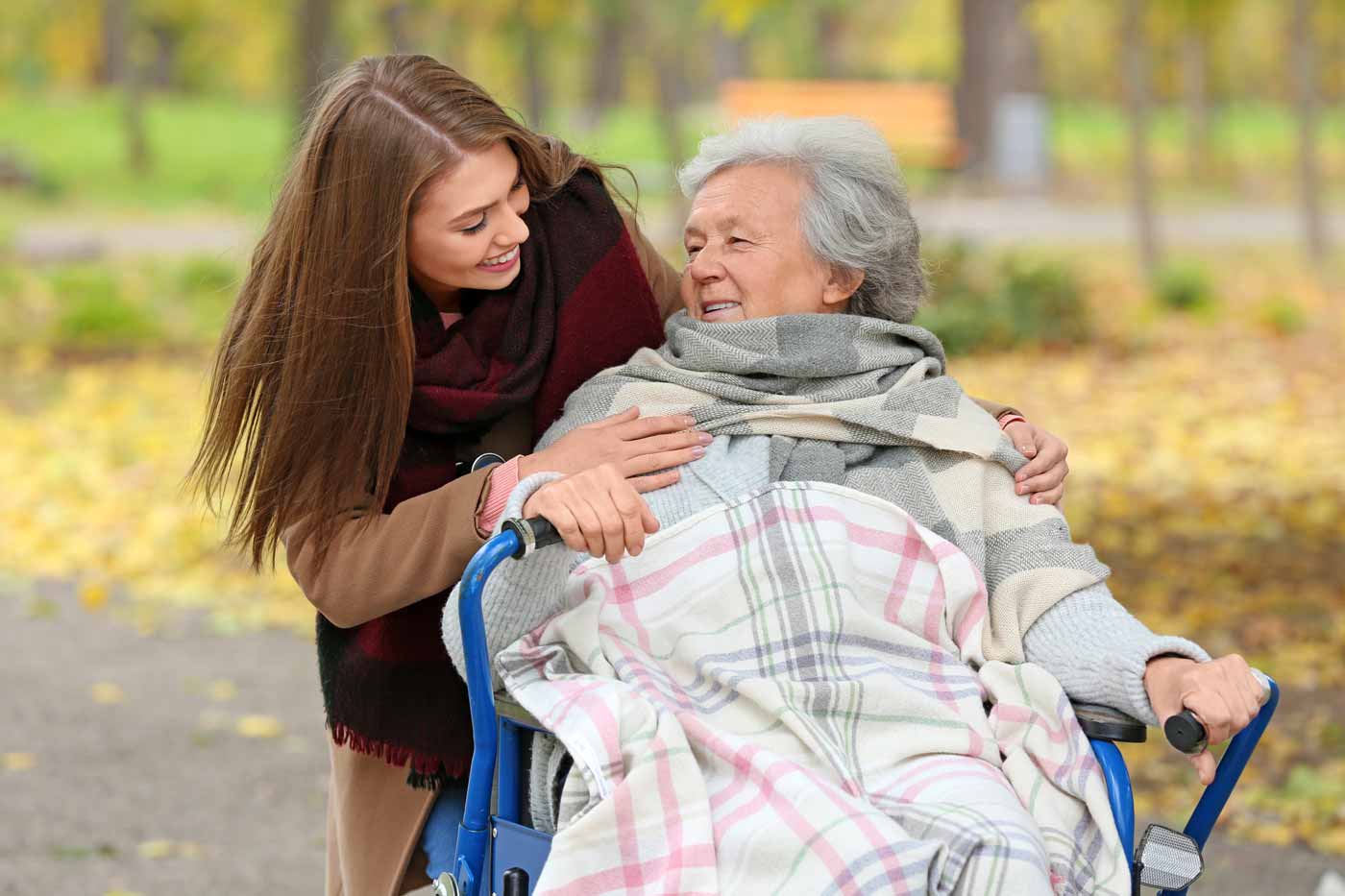A young caregiver smiles and hugs an older woman sitting in a wheelchair, covered with a plaid blanket in an outdoor park with autumn leaves in the background.