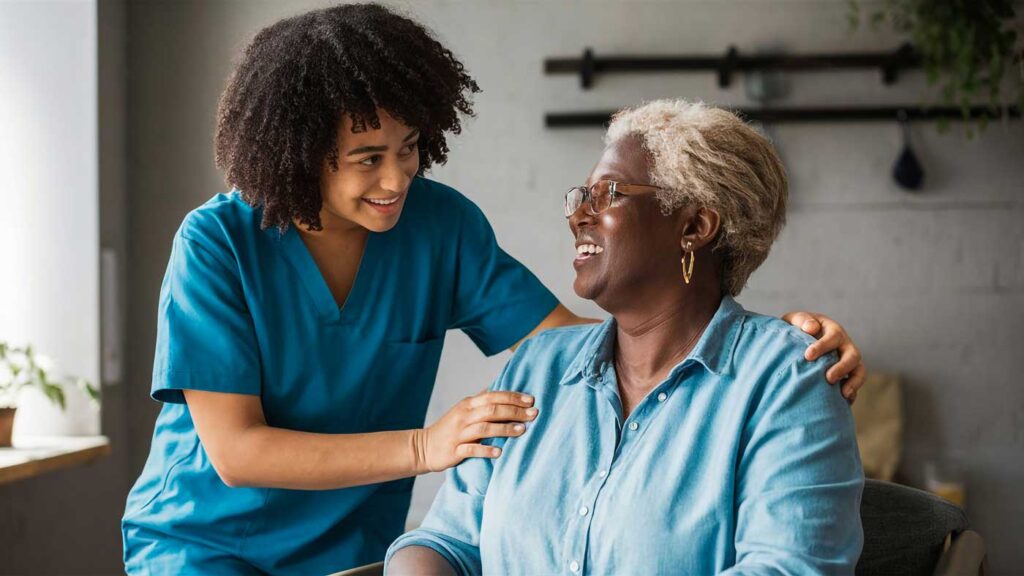 A young healthcare worker in scrubs smiles and gently touches the shoulder of an older woman with gray hair and glasses, who is also smiling. They appear to be in a bright, comfortable room.