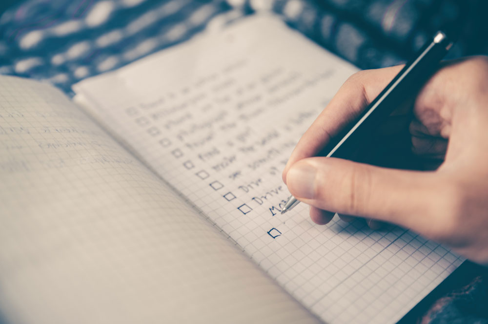 A close-up of a hand holding a pen and writing on a squared notebook with a checklist. Several items are listed, some with boxes checked off and others left blank. The background is softly blurred.