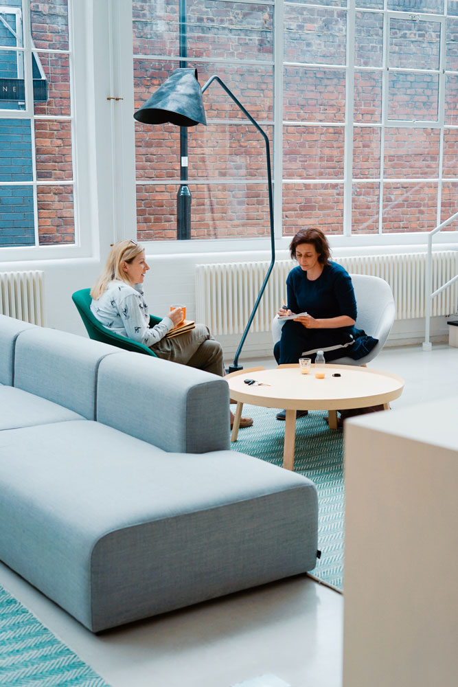 Two women sit across from each other in a modern, bright room with large windows. They are talking and holding drinks at a round wooden table beside a grey sofa and a tall black floor lamp.
