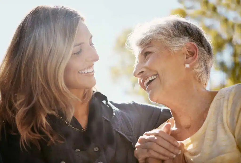 A younger woman and an older woman smile warmly at each other outdoors, holding hands and sharing a close, affectionate moment in the sunlight. Trees are softly blurred in the background.