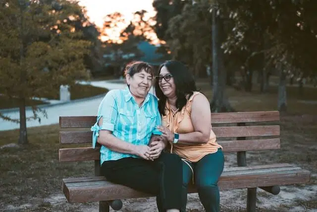 Two women sit closely together on a park bench at sunset, smiling and holding hands. One wears a blue plaid shirt, and the other wears a sleeveless orange top. Trees and a path are visible in the background.