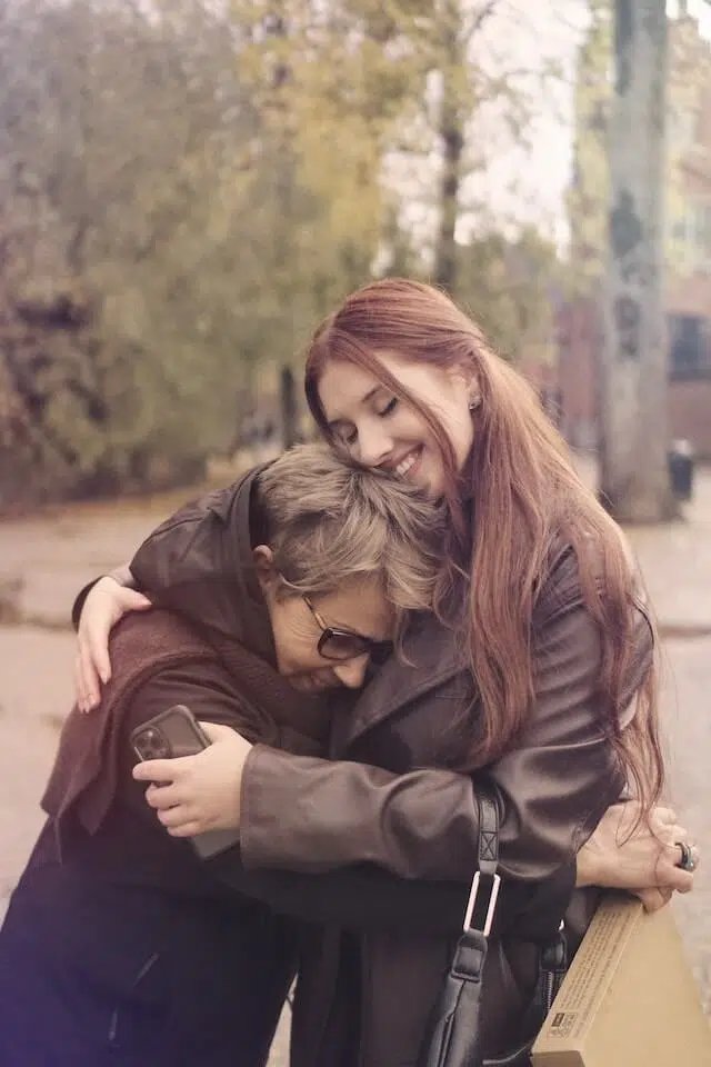 A young woman with long red hair warmly hugs an older woman with short gray hair and sunglasses outdoors on a fall day. Both are smiling, showing affection and closeness. Trees with autumn leaves are in the background.