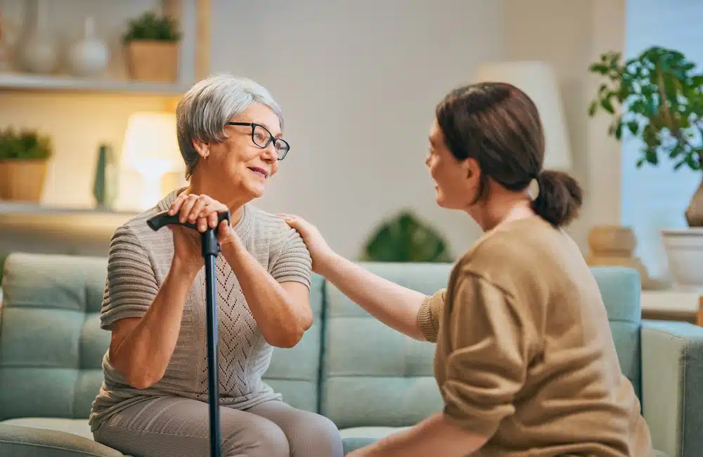 An elderly woman with gray hair and glasses, holding a cane, sits on a couch and smiles warmly at a younger woman who is kneeling beside her, offering comfort and support with a hand on her shoulder.