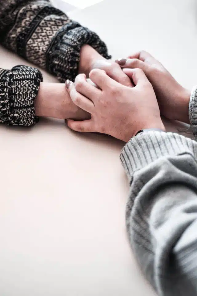 Two people sit across from each other holding hands gently on a table, wearing cozy, patterned sweaters, suggesting comfort, support, and connection.