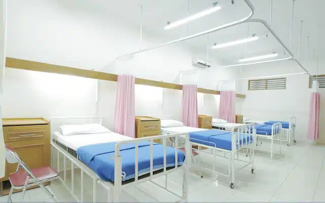 A row of empty hospital beds with blue sheets and white frames, separated by pink curtains, in a bright, clean hospital ward with overhead lighting.