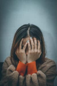 A person wearing a brown sweater with orange sleeves sits against a gray wall, covering their face with both hands, appearing distressed or upset.