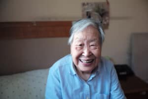 An elderly person with short gray hair smiles warmly while sitting on a bed in a softly lit room, wearing a light blue shirt.