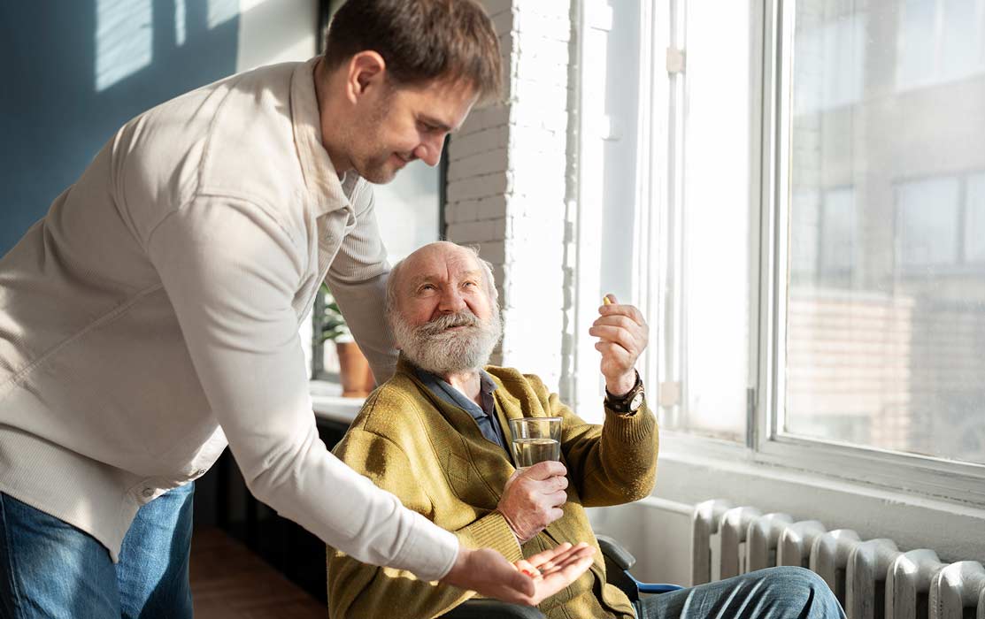 A younger man helps an older man take medication by handing him pills, while the older man sits by a window holding a glass of water and smiling. Sunlight shines into the room.