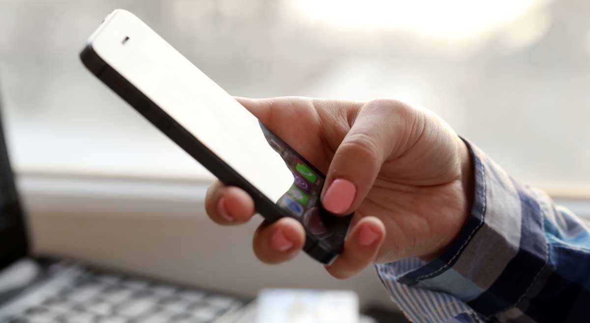 A person with pink nail polish holds a smartphone in their hand, wearing a blue plaid shirt, with a blurred background and sunlight coming through a window.