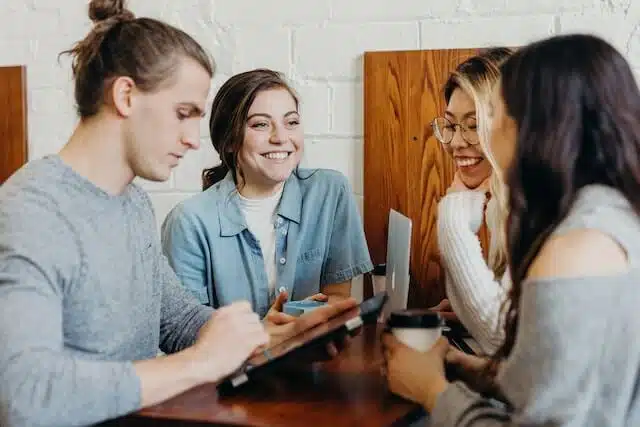 Four young adults sit around a wooden table in a casual setting, talking and smiling. One person writes on a tablet, while others listen and hold drinks, creating a friendly, collaborative atmosphere.