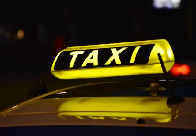 A yellow illuminated taxi sign on the roof of a vehicle at night, with the word TAXI clearly visible in black letters. The background is dark and out of focus.