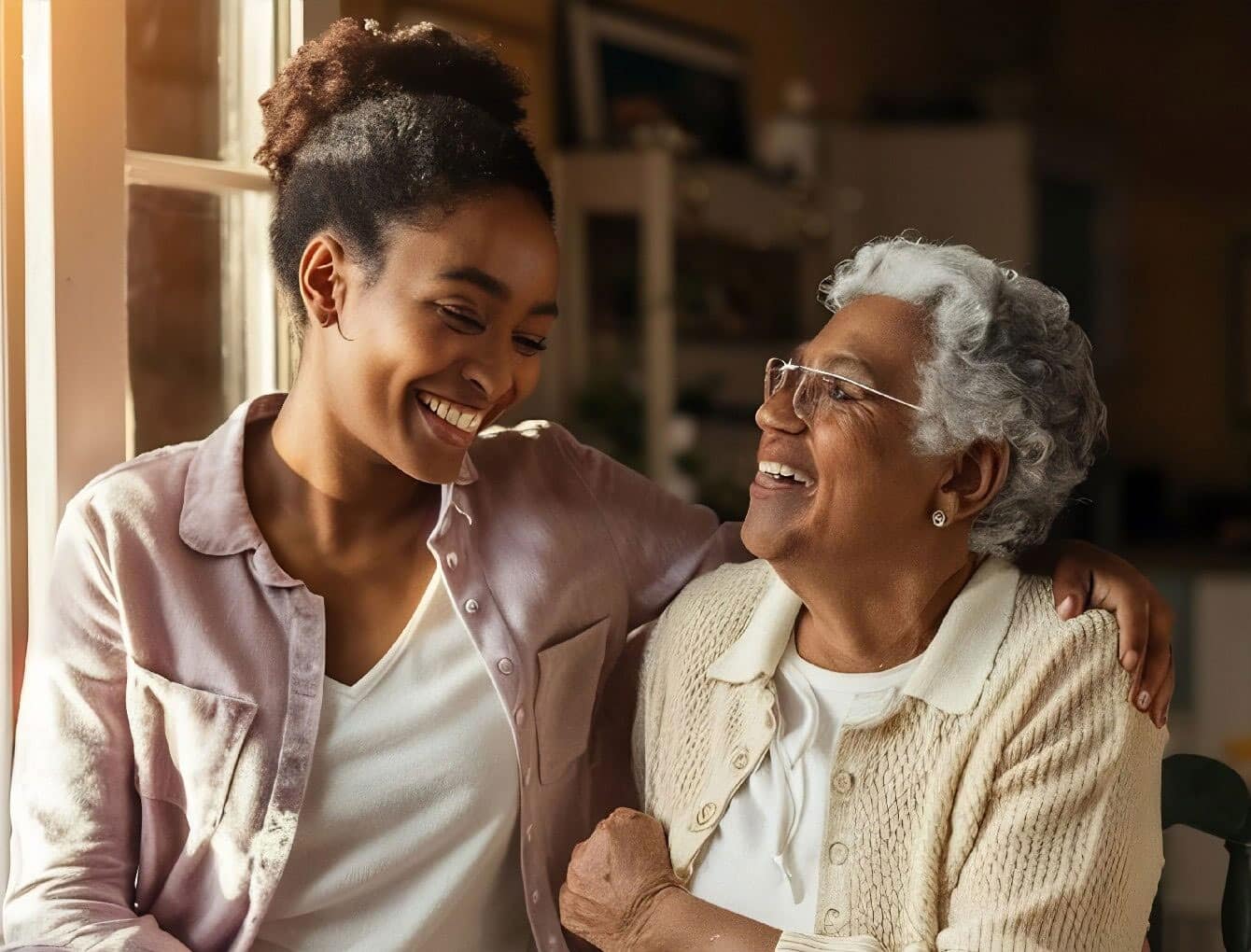 A young woman and an older woman sit closely together by a window, smiling warmly at each other. The younger woman has her arm around the older woman, showing affection and happiness. Sunlight streams in beside them.