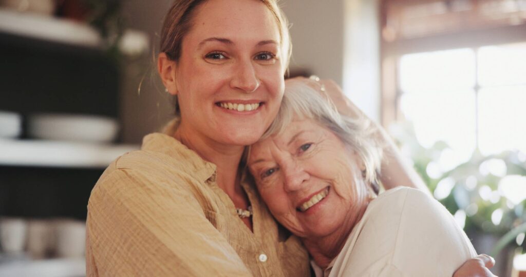 A younger woman and an older woman embrace and smile warmly at the camera in a sunlit room, radiating happiness and affection.