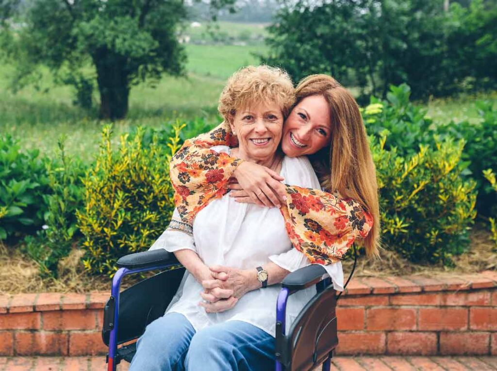 A smiling elderly woman in a wheelchair is embraced from behind by a younger woman outdoors, with greenery and brickwork in the background. Both women look happy and content.