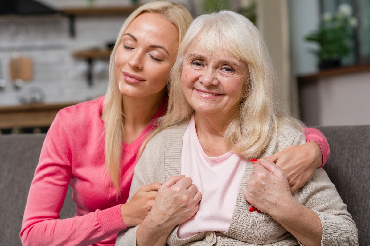 A younger woman hugs an older woman from behind on a sofa. Both are smiling; the older woman looks at the camera while the younger woman has her eyes closed. They appear happy and close, wearing pink and beige sweaters.