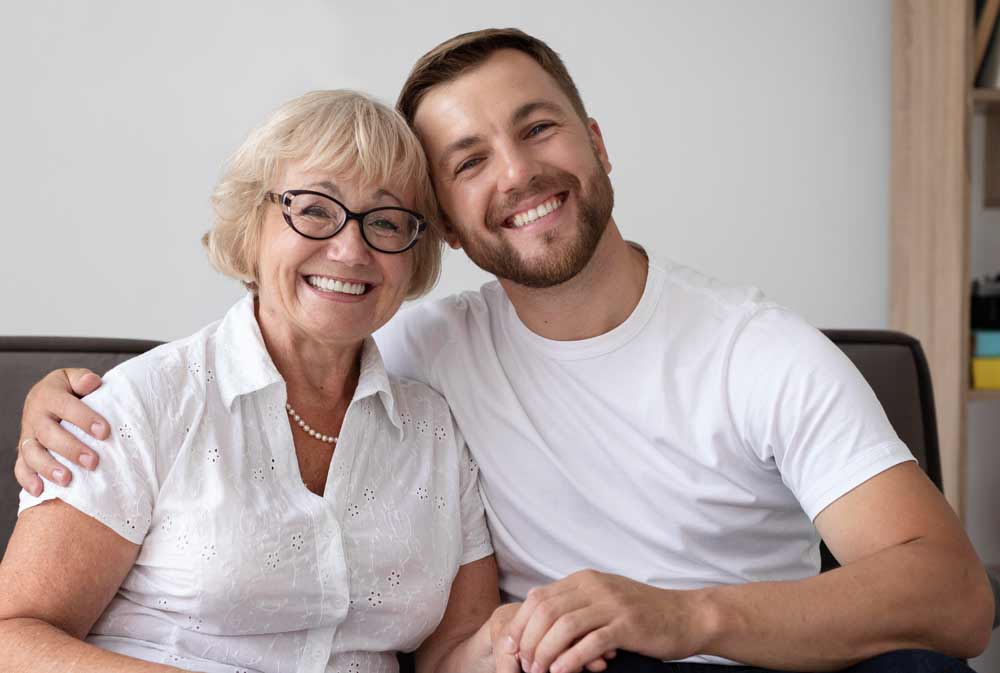 A smiling older woman with glasses and a younger man in white shirts sit close together on a couch, embracing and looking at the camera in a bright, cozy room.
