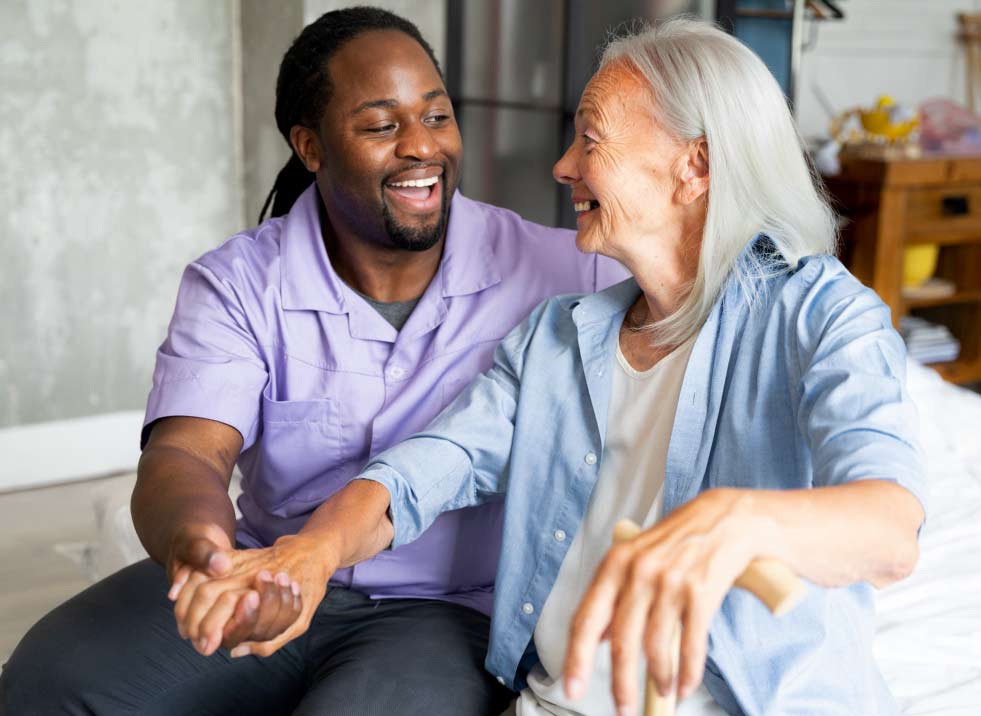 A smiling caregiver holds hands with an elderly woman. They sit close together on a bed, both looking at each other happily, suggesting a warm and supportive relationship.