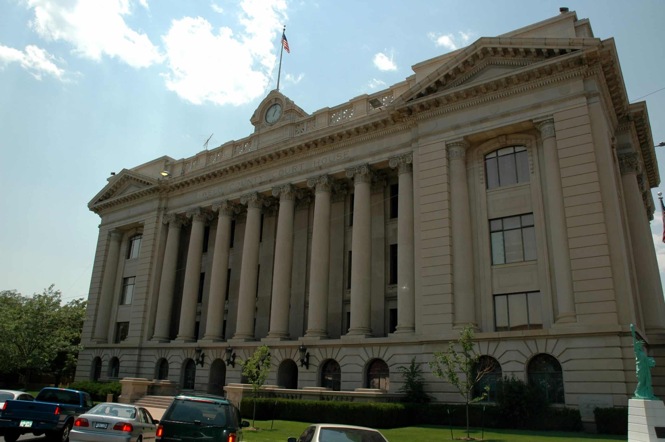 Weld county A large neoclassical courthouse with columns and an American flag on top, cars parked in front, and small trees on the lawn.