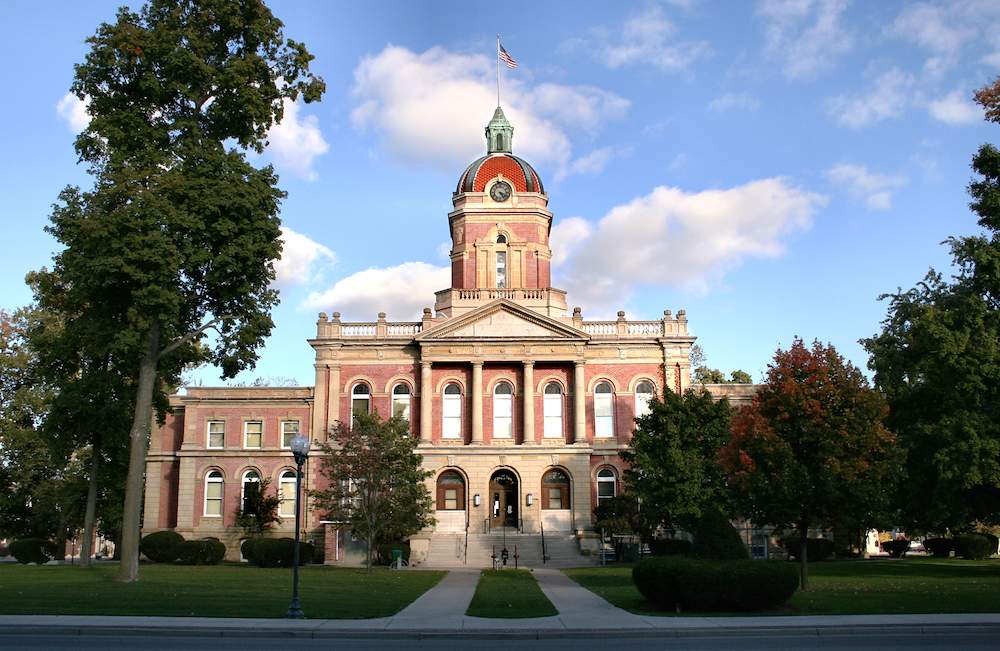 Historic red brick Elkhart County Courthouse on Courthouse Square in downtown Goshen, Indiana, with limestone trim, a central clock tower and dome, and an American flag on a clear day.