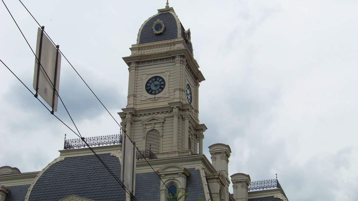 Hamilton County Courthouse clock tower in Noblesville, Indiana, showing the historic cream-colored stone facade, arched windows, Roman numeral clock faces, and mansard roof with decorative iron cresting.