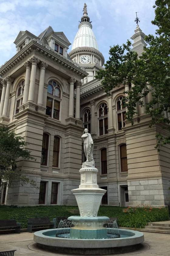 Historic Tippecanoe County Courthouse in Lafayette, Indiana, with its white dome and clock, viewed past a tiered fountain and Lafayette statue on a bright day.