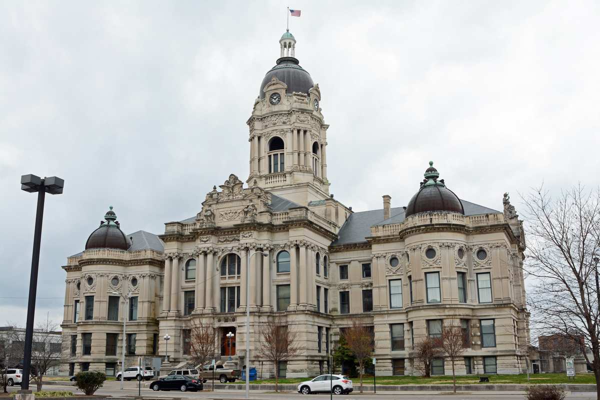 Historic limestone Vanderburgh County Courthouse in Evansville, Indiana, featuring its Beaux-Arts facade, central clock tower with American flag, and multiple dark domes under an overcast sky.