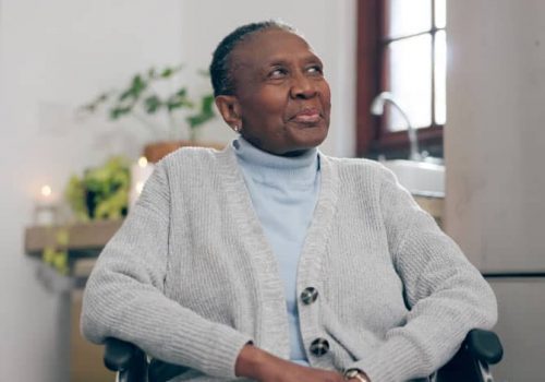 An older woman wearing a light blue turtleneck and a gray cardigan sits indoors, smiling gently and looking slightly upward. A plant and a window are visible in the softly lit background.